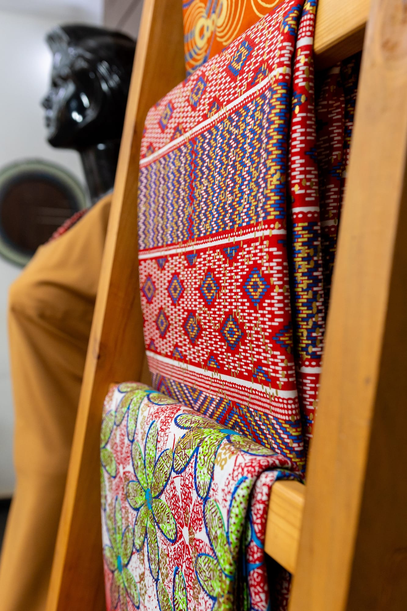 Bold African wax-print fabrics draped over a wooden ladder display beside a carved bust in the boutique