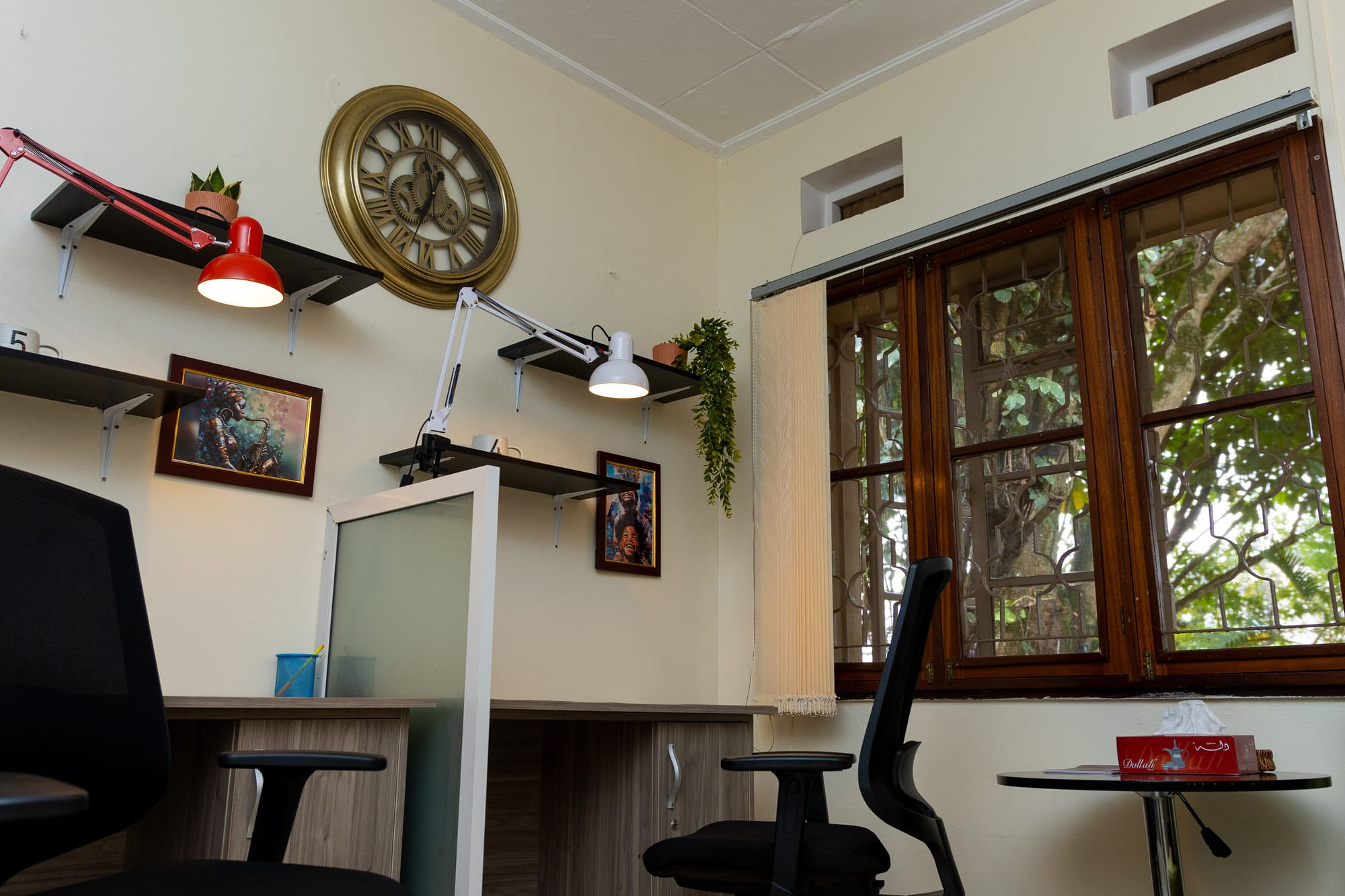 A coworking desk by the window at KaraSpace — adjustable lamps, a vintage clock, framed jazz art and a leafy view through the trees