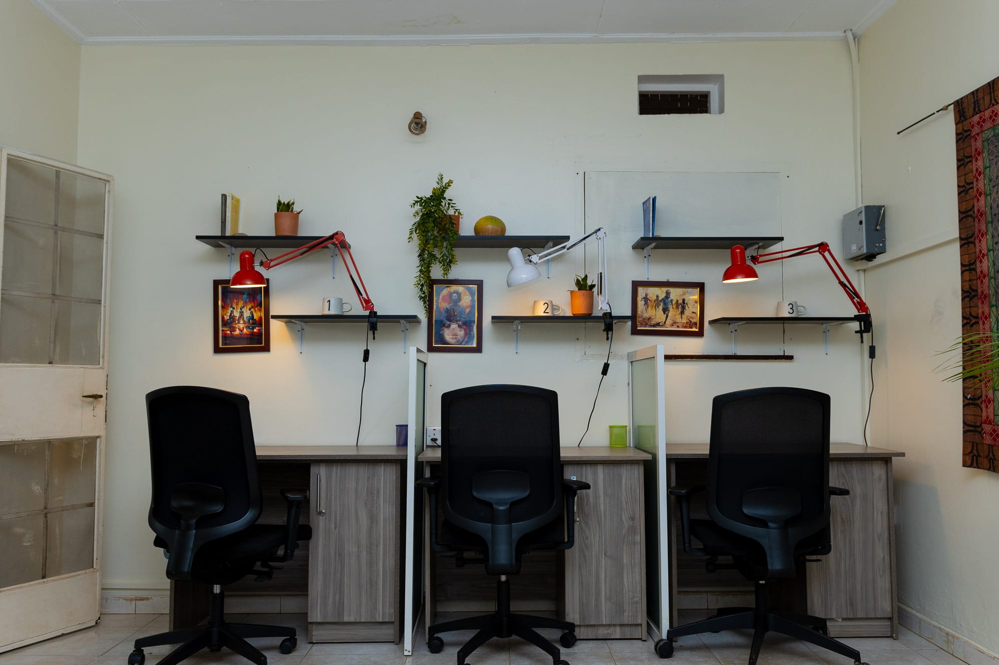 A row of three coworking desks at KaraSpace with black ergonomic chairs, adjustable lamps and framed African art on the shelves