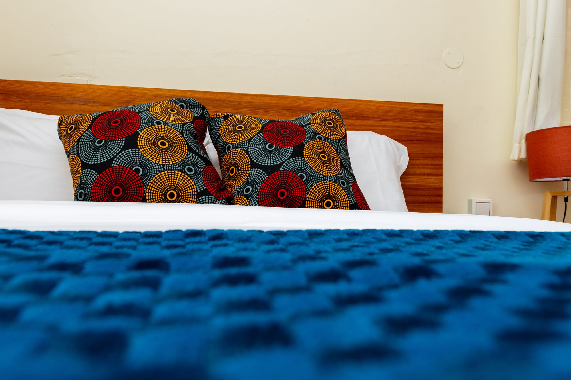 Deluxe Room — a double bed with blue throw and African print cushions beside a bright window