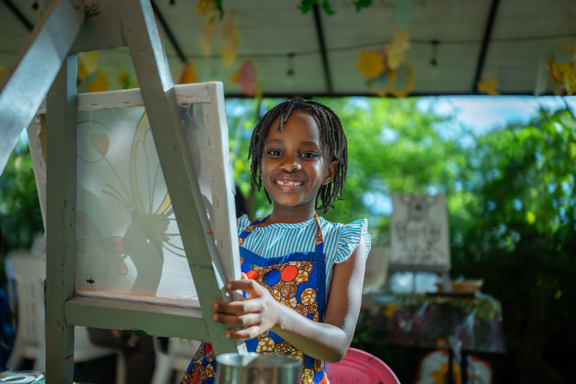A young maker in an African print apron beaming beside her easel